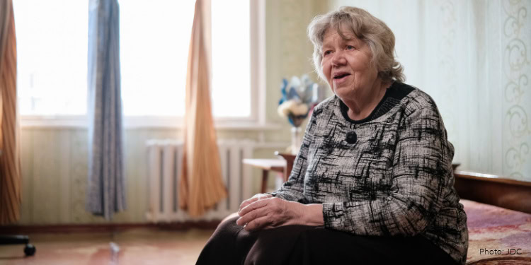 Supportive elderly woman sitting on a sofa, speaking and smiling in a cozy living room with natural light, representing community support and care for seniors.