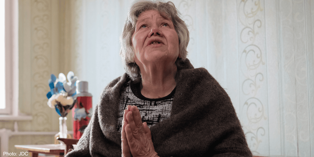 Elderly woman with gray hair, eyes closed, hands clasped in prayer, seeking help or comfort, in a cozy home setting.