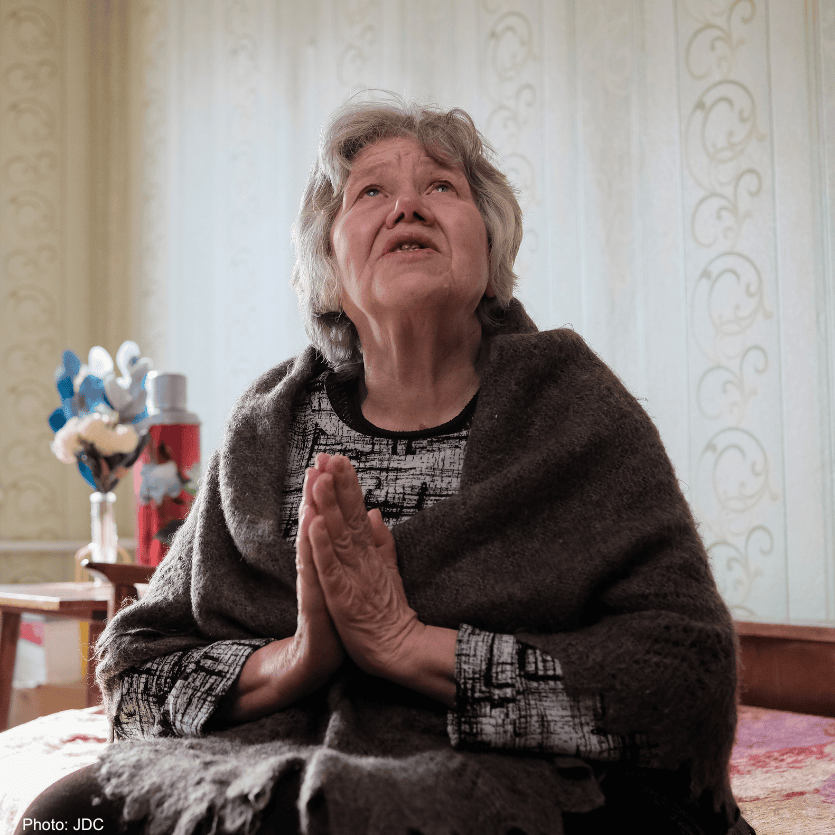 Elderly woman with gray hair, eyes closed, hands clasped in prayer, seeking help or comfort, in a cozy home setting.