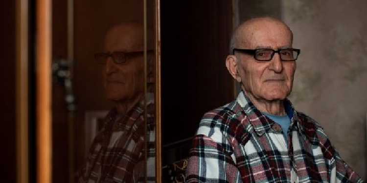 Elderly Jewish man sitting next to a mirror looking directly into the camera.