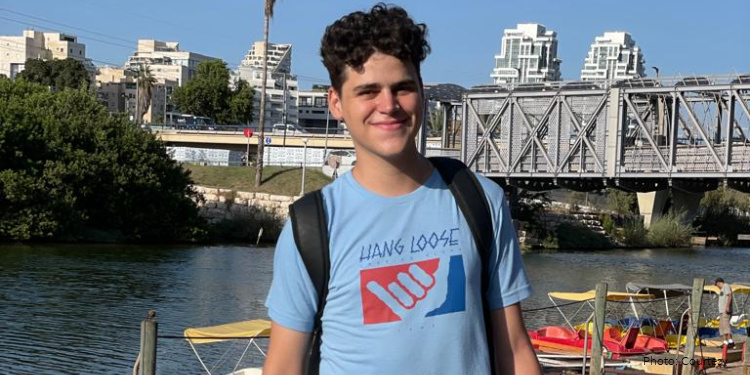 Matias Golda, oleh from Argentina. young adult man standing near river outdoors, boats by dock, bridge, light blue shirt and gray shorts, text and graphic on shirt reading "hang loose", smiling, happy