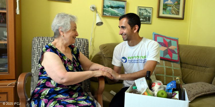 An elderly person seated in a chair holding hands with a younger individual wearing a white t-shirt, with a box of groceries including fruits placed in front of them. The background features framed pictures and home furniture, suggesting a warm, domestic setting.