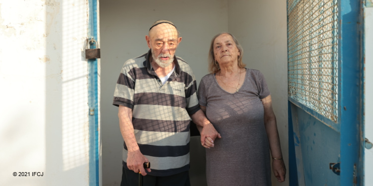 Elderly couple stand in the doorway holding hands.
