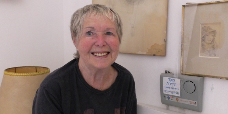 Miriam, IFCJ recipient smiling while standing in her home with old paintings and an IFCJ sponsored emergency call button.