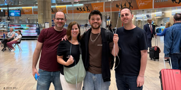 Nathan Israel - Aliyah from Chile - October 22, 2025 group of 4 people at an airport - holding large balloons, gold balloons, Israel balloon - man with glasses, maroon T-shirt, sandals - woman, black shirt, green purse - young man, beard, brown t-shirt, gold chain - man, black T-shirt, jeans