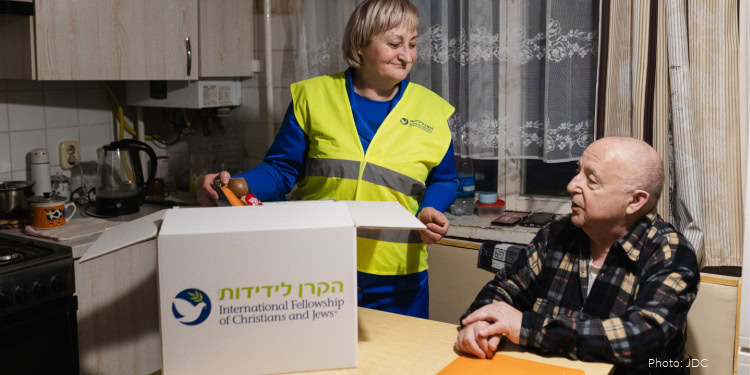 Visit to Nicolay Bogulavskiy - Kishinev, Moldova - February 12, 2026 blonde woman, blue shirt, branded logo safety vest - opening branded logo food box on kitchen table - food items on table - elderly man, blue and cream plaid shirt - sitting at kitchen table - windows, white lace curtains