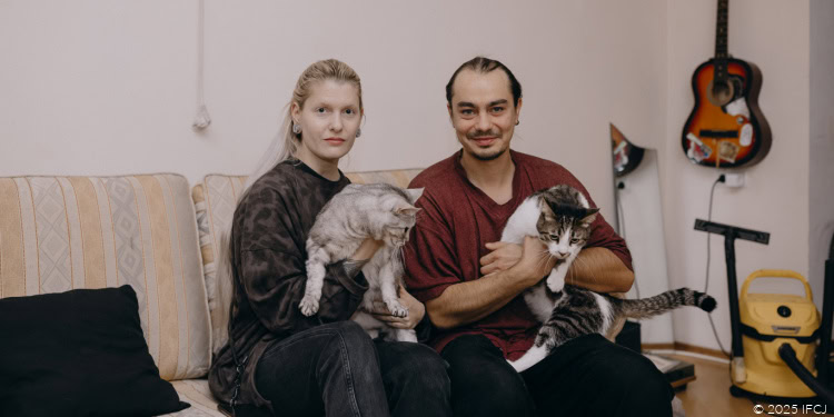 Nikita Poloskov and Alexandra Burenkova - Russian Aliyah - Tbilisi, Georgia - October 20, 2025 blonde woman holding a light gray cat - man in maroon t-shirt holding a dark gray and white cat with stripes - sitting on a light grey and light orange striped couch - guitar on wall - yellow vacuum cleaner - looking at camera