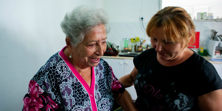 Elderly woman in pink shirt smiling while a woman in a black shirt is helping her.