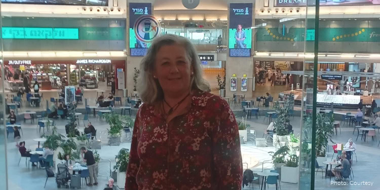 A woman with gray hair smiling at the camera in an airport terminal, surrounded by travelers and seating areas, representing IFCJ's efforts to assist those in need worldwide.