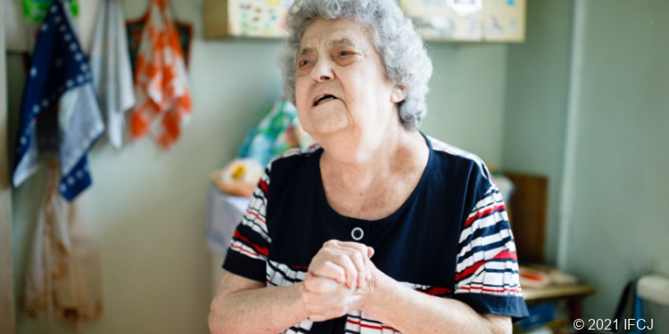 Elderly woman standing in kitchen with hands folded together