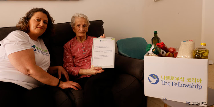Two women, one elderly and one younger, sitting on a couch with a box of supplies and a document, representing community support and aid.
