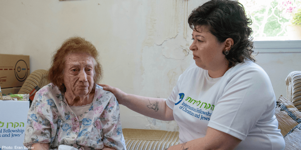 Nataly Tilkin (IFCJ Staff) sitting with elderly woman, pink floral dress - white branded branded logo T-shirt, brown, tan couch - Nataly's hand on her shoulder, looking at each her, listening, caring - branded logo food box in rear