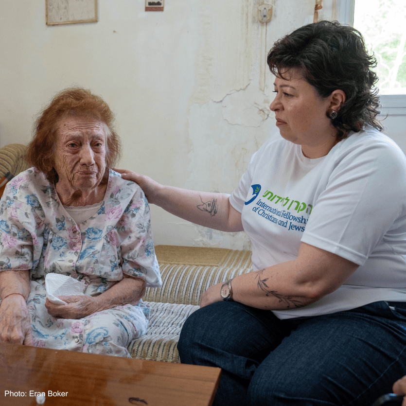 Nataly Tilkin (IFCJ Staff) sitting with elderly woman, pink floral dress - white branded branded logo T-shirt, brown, tan couch - Nataly's hand on her shoulder, looking at each her, listening, caring - branded logo food box in rear