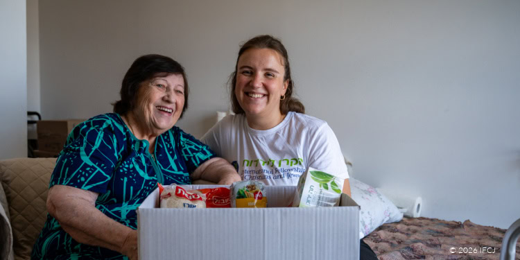 Duas mulheres, uma idosa e outra jovem, sorridentes e sentadas juntas ao lado de uma caixa de ajuda alimentar, simbolizando o apoio da comunidade e o trabalho de caridade.