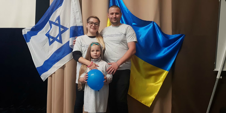 Smiling family of three standing in front of two flags.