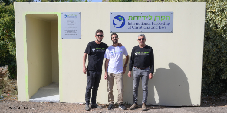 Three men in IFCJ branded shirts standing outside an IFCJ branded bomb shelter funded by donors Robert Steil and Janet Steil.