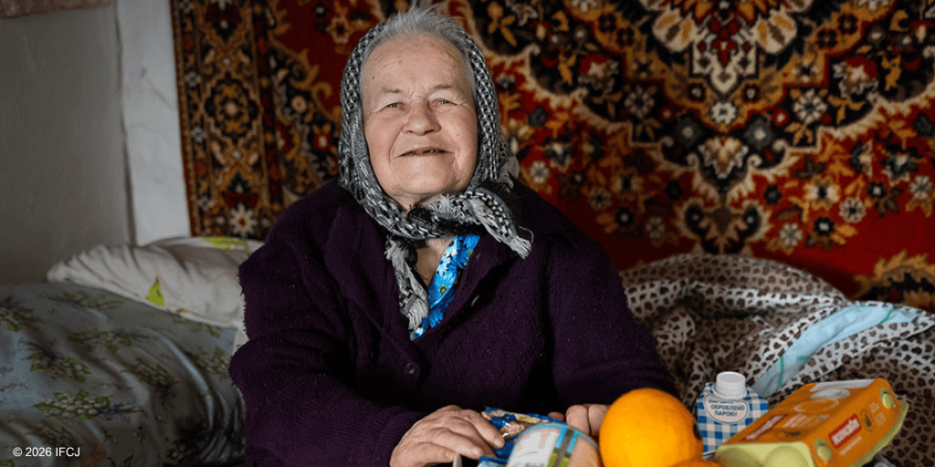 Smiling elderly woman with gray hair and headscarf sitting on a bed, surrounded by colorful textiles and personal care items, in a warm home setting.