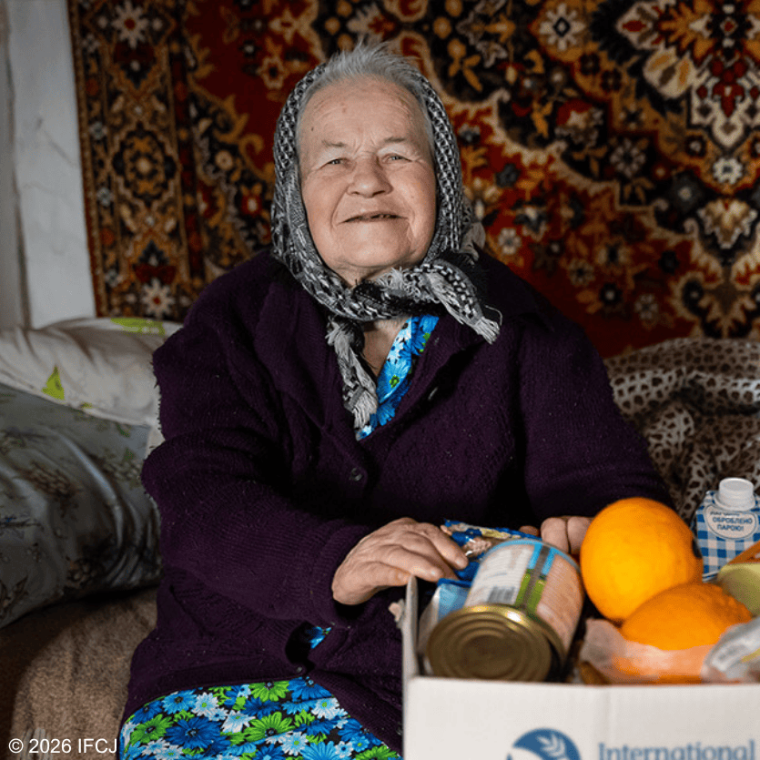 Smiling elderly woman with gray hair and headscarf sitting on a bed, surrounded by colorful textiles and personal care items, in a warm home setting.
