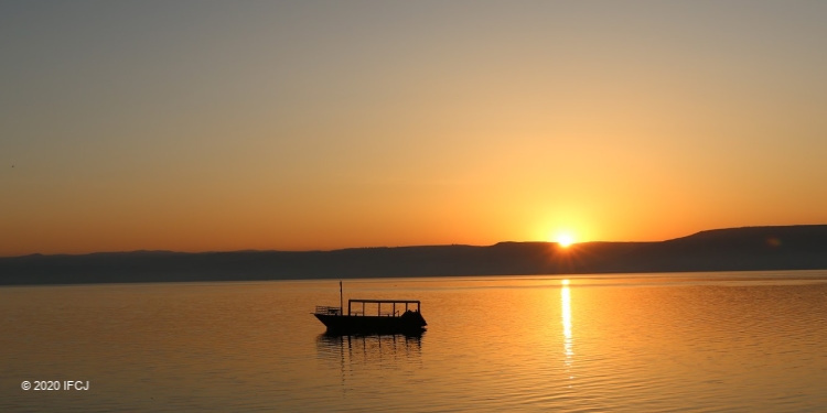 The sun rising over the Sea of Galilee as a single boat is in the water.