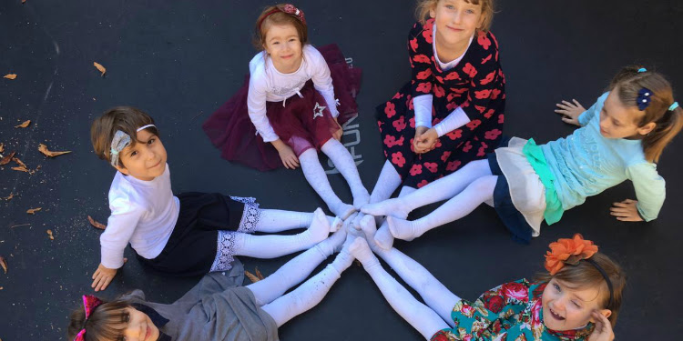 Six young girls sitting on the sidewalk together smiling up at the camera.