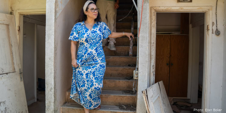A woman wearing a blue and white patterned dress and glasses stands on a staircase inside a home, holding the railing and looking to the side.