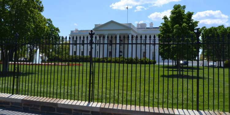 The White House, the official residence of the U.S. President, is shown behind a black metal fence with green trees and a blue sky in the background.