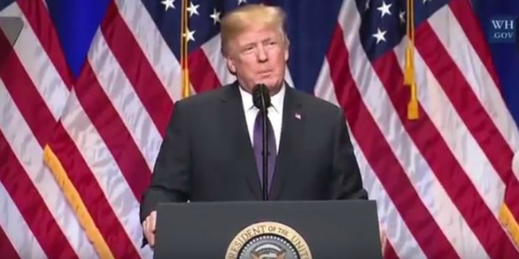 President Trump giving a speech at a podium with three American flags behind him.