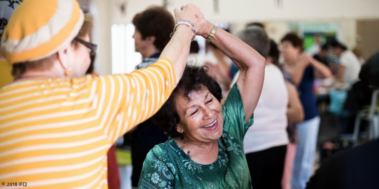 An elderly woman in yellow twirling an elderly woman wearing green while dancing.