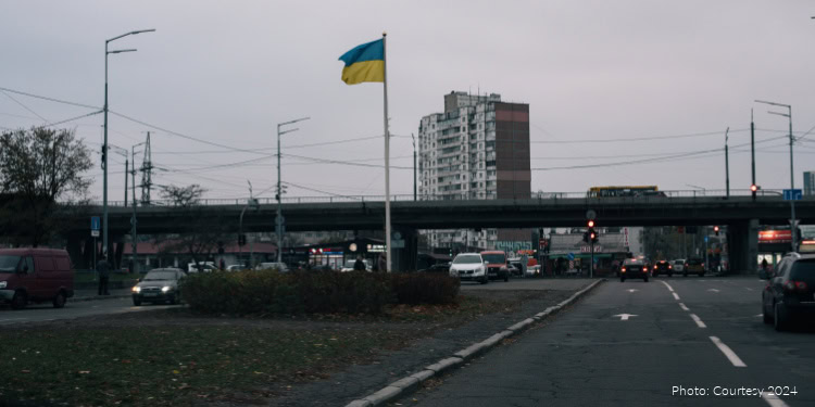 Ukrainian flag flying high over a busy city street with cars and a bridge, under an overcast sky, symbolizing hope and resilience.