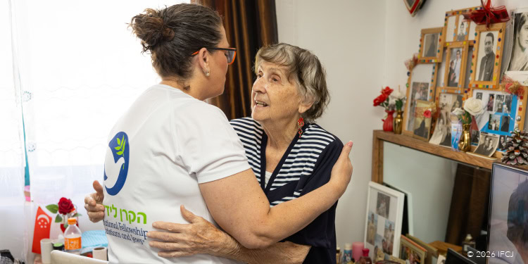 Elderly woman with gray hair and striped shirt being comforted by a caregiver in a cozy home setting in Jerusalem.