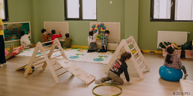 Visit to Or Avner FJC Jewish School - Tbilisi, Georgia (FSU) - October 20, 2025 playroom for young children - light green walls, windows, black frames, blinds - children playing - light wood climbing ladders - rug with trees on it - children putting pegs on a wall board - green hoop - boy in his knees, black shirt with red numbers on back