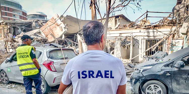 Man in a white shirt with Israel on the back of it staring at the destruction of a building.
