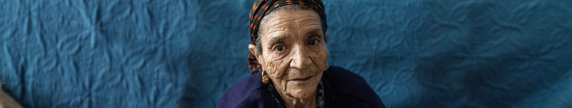 Elderly Jewish woman sits on couch wearing scarf on her head and looking sad