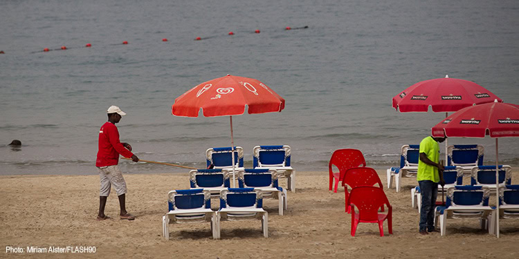 Beach scene with red umbrellas, blue and red chairs, and people setting up on the sandy shore near calm water.