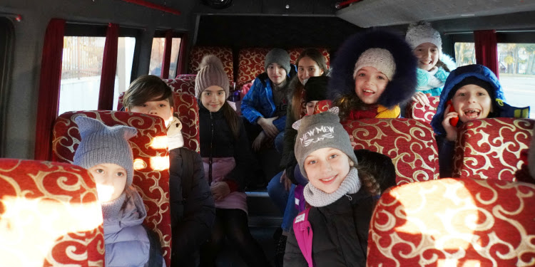 Young girls sitting on a bus with red seats smiling at the camera.