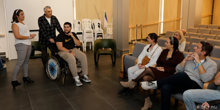 Davida Kutscher (IFCJ Staff), white blouse, white headband, standing to left - woundd soldier sitting in wheelchair, T-shirt with Nike logo - man in plaid shirt standing with hands on wheelchair - group of people sitting in auditorium seats - Yael Eckstein, dark brown dress, Clay Travis in light gray sweater Sheba Medical Center (aka Sheba Tel HaShomer hospital) - The Fellowship has funded advanced rehabilitation equipment and the expansion and renovation of the physical therapy facilities within the Head Injury Rehabilitation Department. Clay and IFCJ staff met with wounded soldiers who are receiving Fellowship support and distributed gifts.
