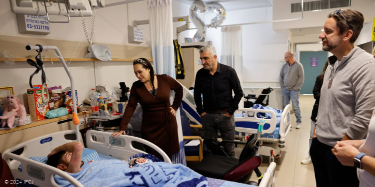 Clay Travis Tour of Israel - Sheba Medical Center - Ramat Gan - December 10, 2024 Yael Eckstein, dark brown dress, Clay Travis, light gray sweater, and Safwan Marich (IFCJ Staff) visiting wounded soldier laying in hospital bed - young adult man with light blue blanket Ariel Kokashvili lost both legs and sustained additional severe injuries while clearing ordinance from a home in Gaza Sheba Medical Center (aka Sheba Tel HaShomer hospital) - The Fellowship has funded advanced rehabilitation equipment and the expansion and renovation of the physical therapy facilities within the Head Injury Rehabilitation Department. Clay and IFCJ staff met with wounded soldiers who are receiving Fellowship support and distributed gifts.