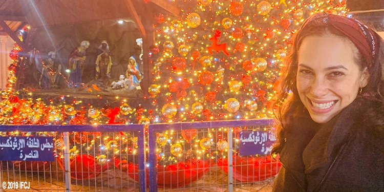Yael Eckstein smiling next to a lit Christmas tree and the nativity scene.