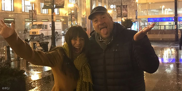 Two people smiling and celebrating in the rain at night in an urban setting, with reflections on wet streets and city lights in the background.