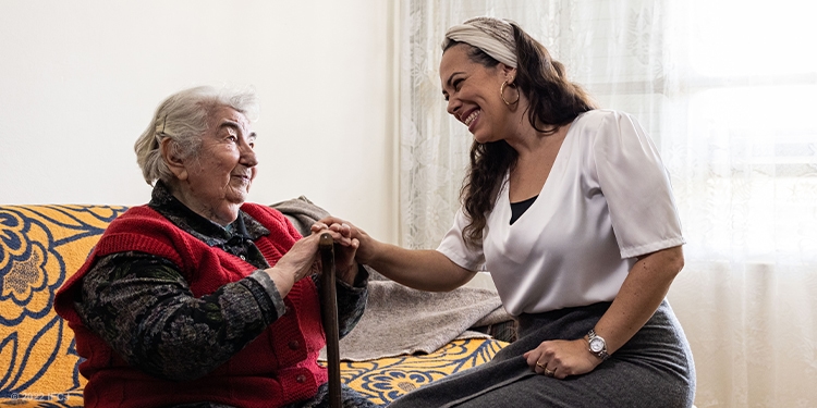 Yael smiling at an elderly Jewish woman while they sit together on a yellow couch.