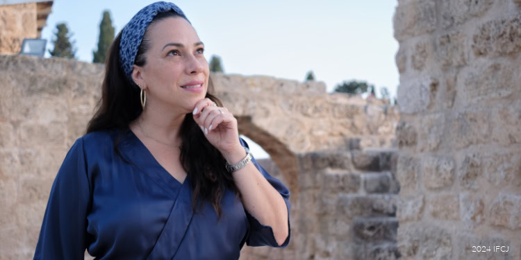 Woman in blue attire with headband, looking thoughtfully into the distance against a historic stone backdrop, symbolizing hope and resilience in Israel.