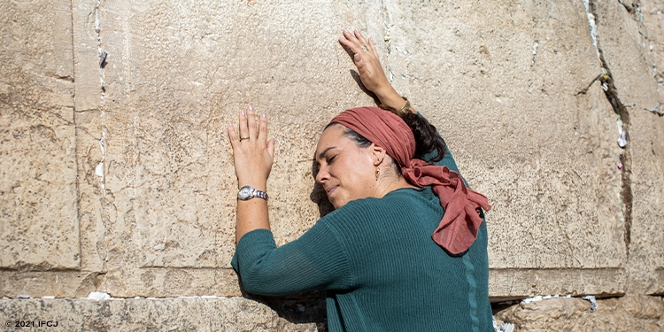 Yael Eckstein, wearing a green shirt and red head covering, leans against the Western Wall in prayer.