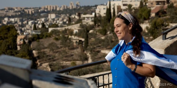 Yael in Jerusalem - October 20, 2025 Yael Eckstein - bright blue dress, tan and white headscarf, smiling, Israeli flag around her shoulders, blowing in wind - standing on a balcony, metal railing - looking at the view - scenic view