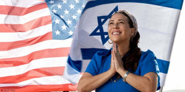 Yael in Jerusalem - October 20, 2025 Yael Eckstein - bright blue dress, tan and white headscarf - standing in front of Israeli flag and American flag, blowing in wind - eyes open, smiling, head up, hands together in prayer