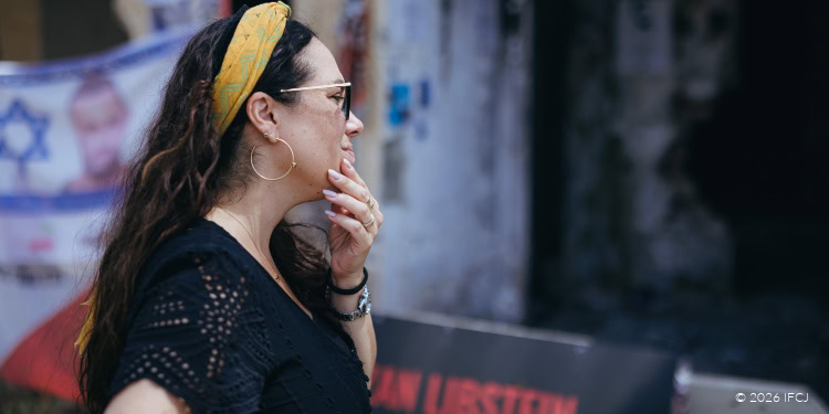 A woman wearing sunglasses and a yellow headband, standing in front of a protest banner, appears contemplative and concerned.
