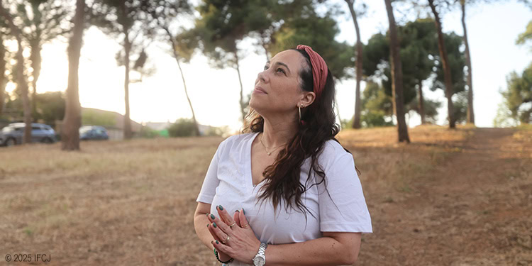 Yael standing outdoors by trees looking up at the sky with praying hands