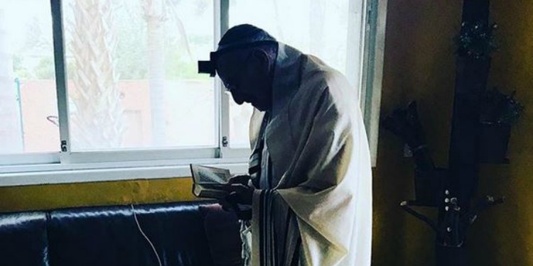 Elderly man in white robe looking down at book beside window.