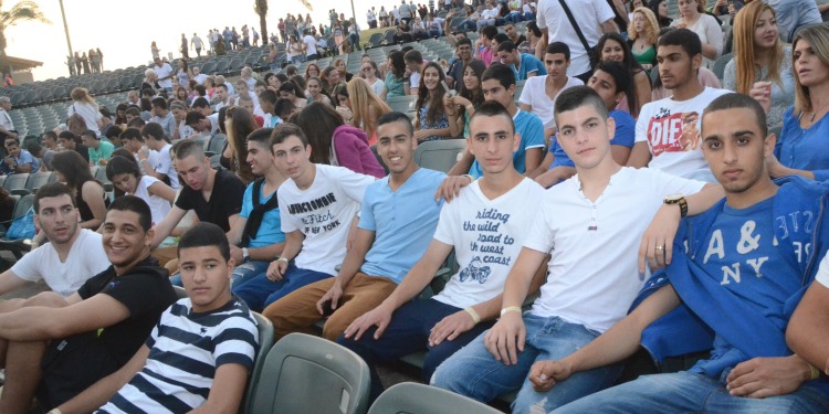 Group of several teenage boys sitting in green bleachers in a crowd.