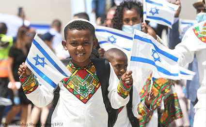 Ethiopian aliyah, June 1, 2022, group of olim on tarmac, boy waving flags of Israel, other passengers out of focus in the background, white shirt, traditional clothing, yarmulke.