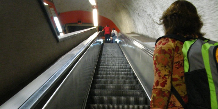 The back of a young woman going up an escalator.